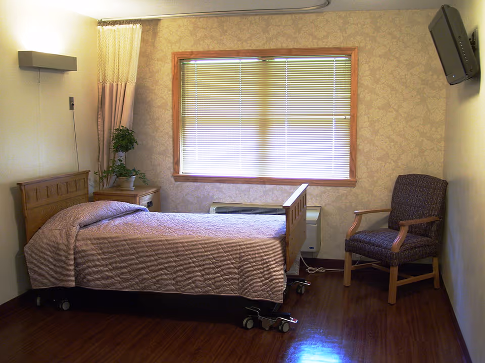 A small nursing facility bedroom with a single bed covered in a light purple quilt, a wooden nightstand with a potted plant, a window with closed blinds, a patterned armchair, and a wall-mounted television.