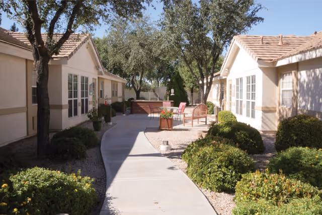 Outdoor walkway between two single-story buildings with beige walls and tiled roofs, surrounded by green bushes and trees, with patio furniture including chairs and a table in the background under clear blue sky.