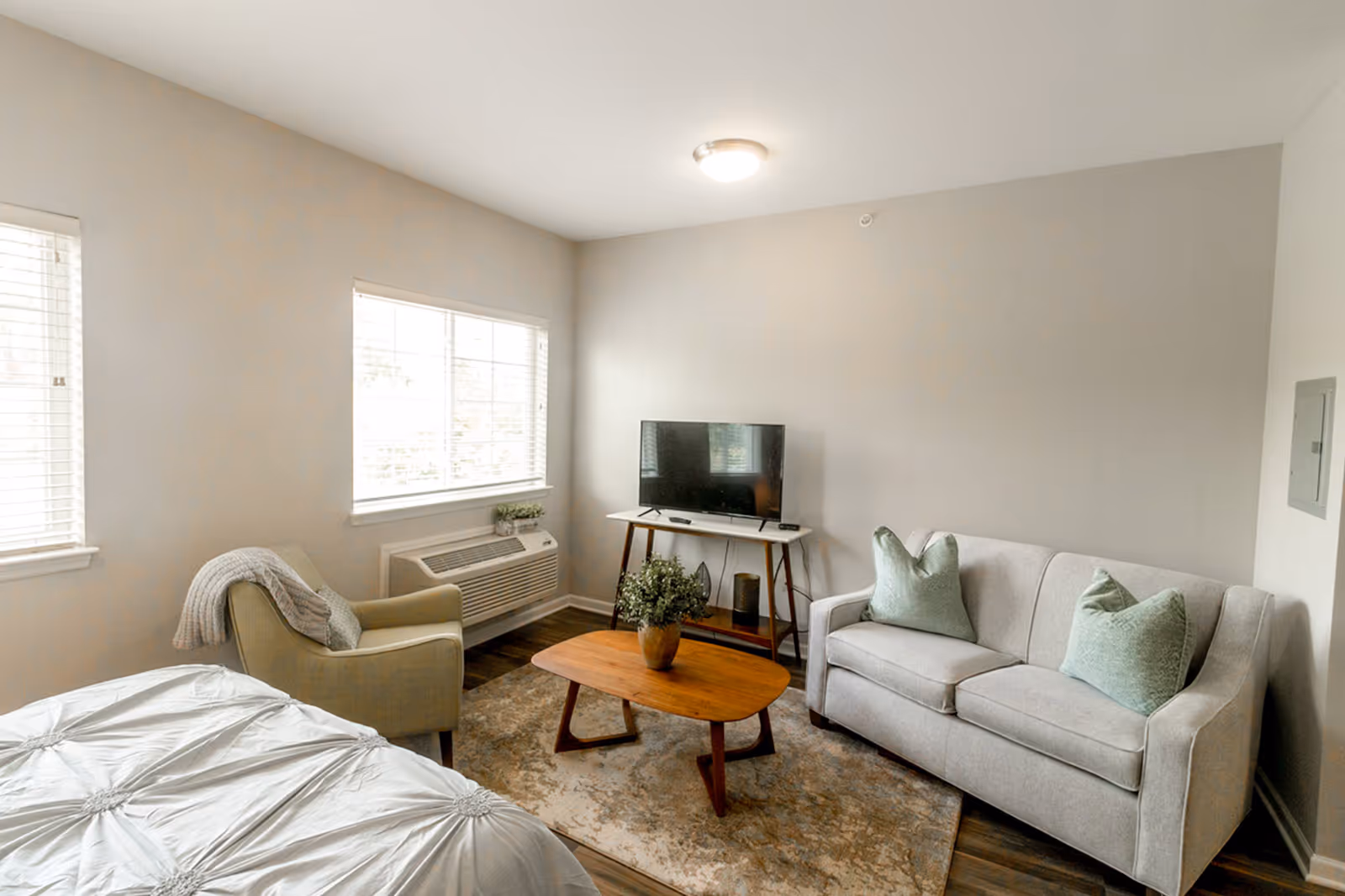 Bright studio living area with a sofa, armchair, coffee table, TV, and a bed near two windows.