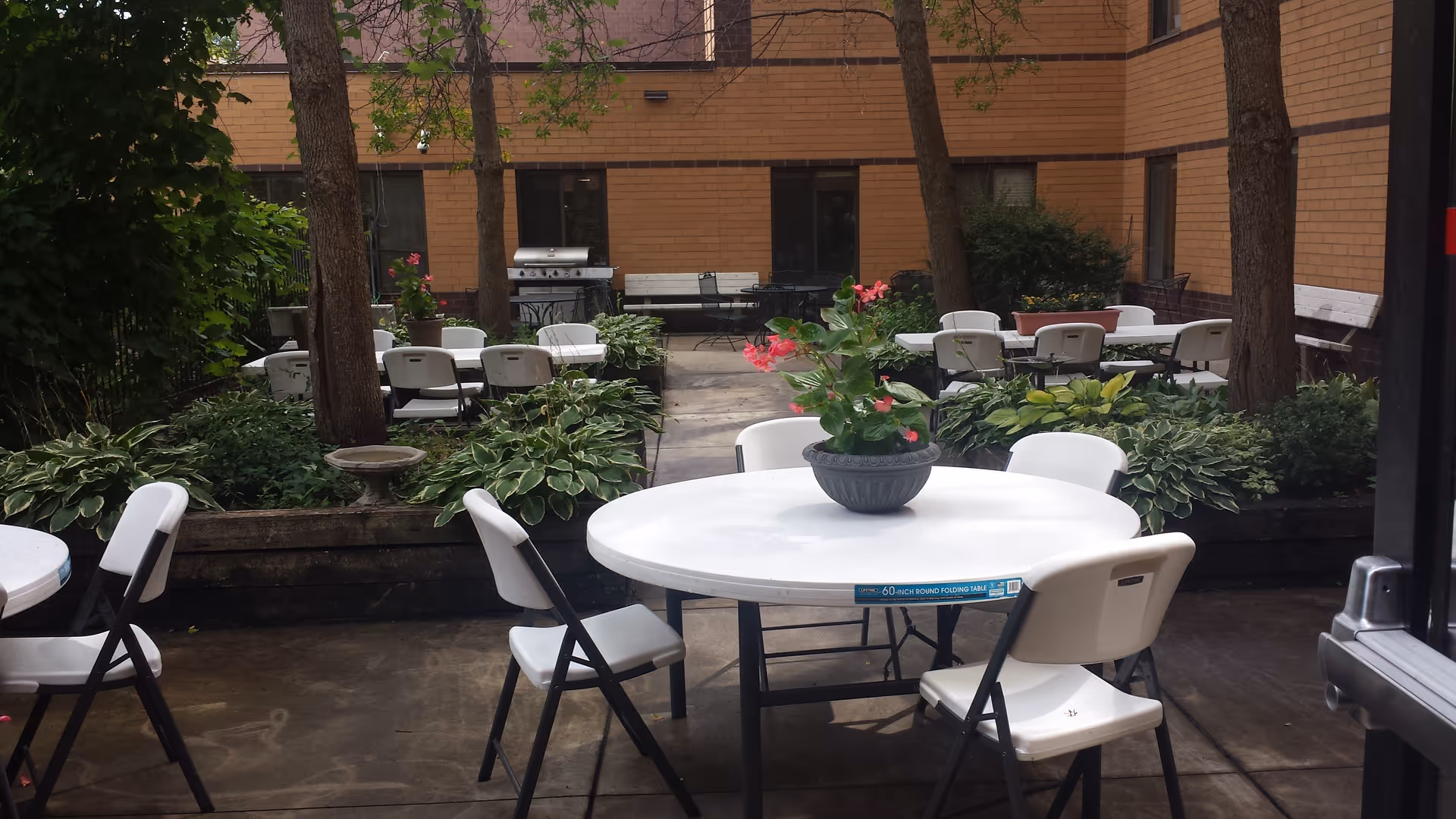 Courtyard seating area with white round folding tables and chairs, potted flowers, and surrounding brick building.
