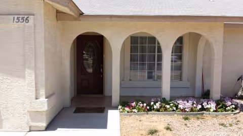 Front entrance of a stucco senior living facility with arched walkway and blooming flower bed.