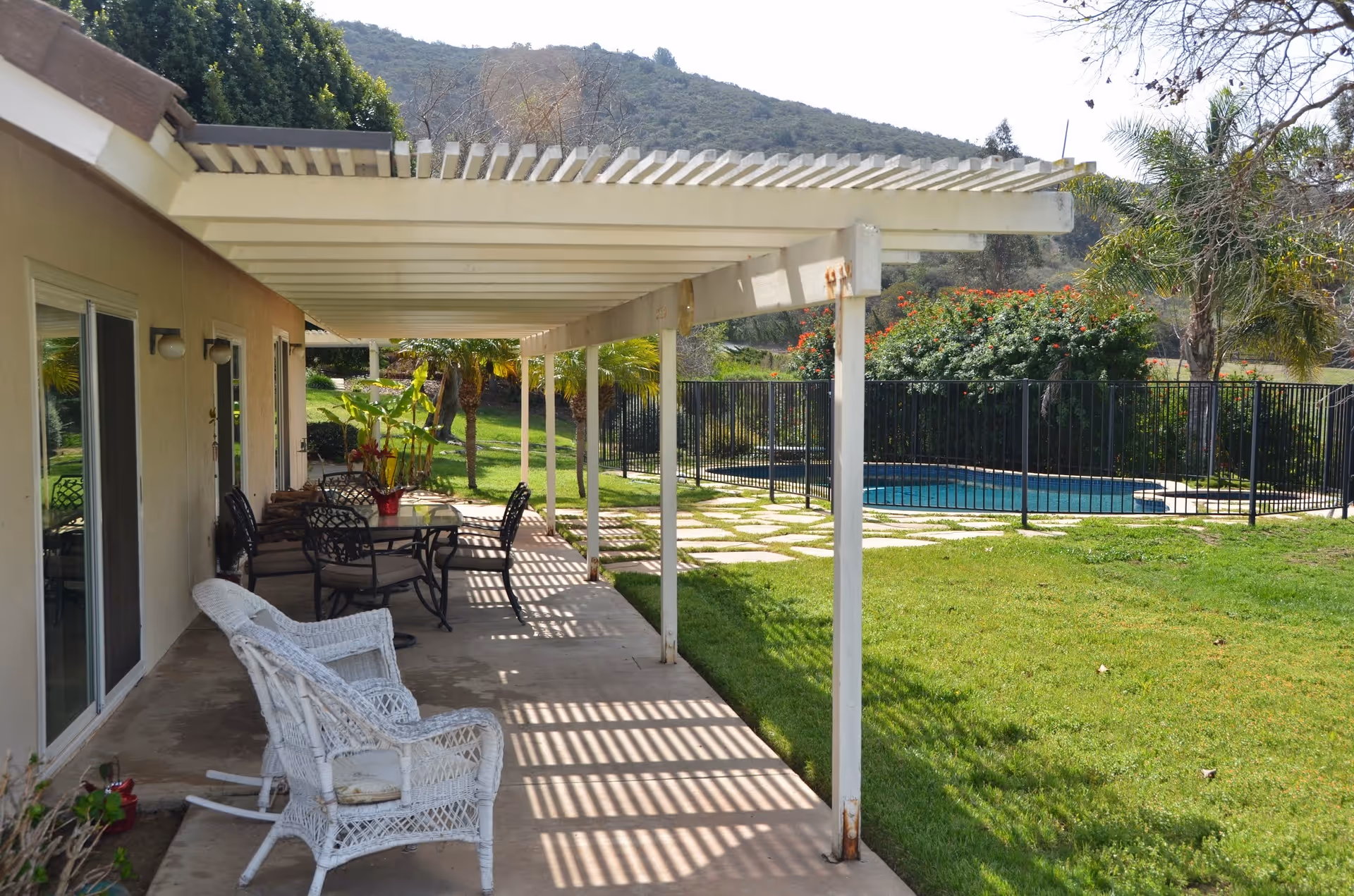 Outdoor patio area at Daybreak Retirement Villa with a white pergola providing shade over a concrete walkway. There are white wicker chairs and a glass-top table with black metal chairs. In the background, there is a fenced swimming pool, green lawn, palm trees, and hills.
