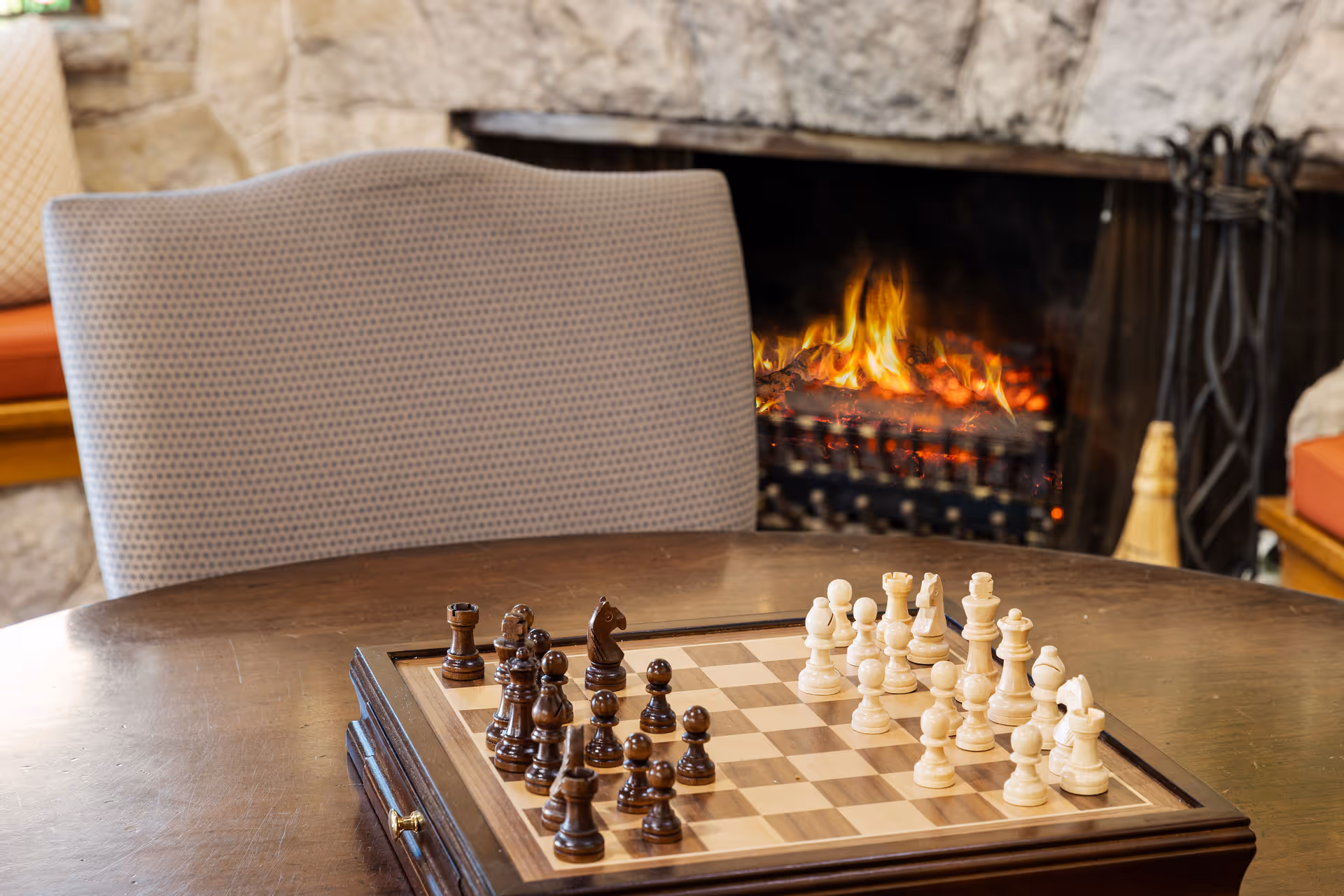A chessboard with wooden chess pieces set up on a wooden table in front of a lit fireplace. A cushioned chair is positioned behind the table, and the background features a stone wall and fireplace tools.