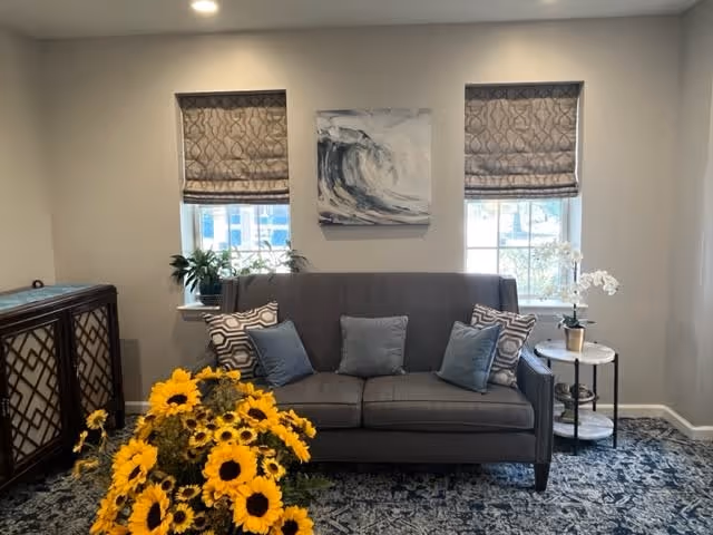 A cozy living room area with a gray sofa adorned with five decorative pillows in shades of blue and gray. Behind the sofa are two windows with patterned Roman shades and a painting of a wave centered between them. To the left of the sofa is a wooden cabinet with a lattice design, and to the right is a small round side table with a white orchid plant. In the foreground, there is a large bouquet of bright yellow sunflowers. The room has light-colored walls and a patterned blue carpet.