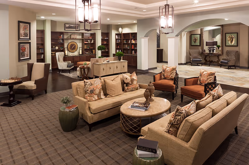 A spacious and elegantly decorated living room area with beige sofas adorned with patterned cushions, two orange armchairs, a round coffee table with decorative items, and a side table with books. The background features built-in wooden bookshelves filled with books and decorative pieces, a small seating area with a wingback chair, and a mirrored wall with two patterned chairs and a table with a lamp. The room has warm lighting from hanging pendant lights and recessed ceiling lights.