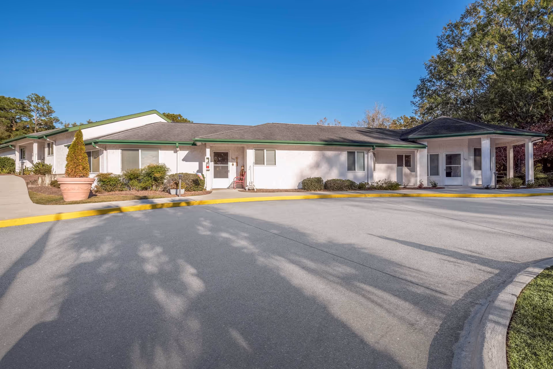 Exterior view of a single-story building with white walls and a gray roof with green trim, surrounded by shrubs and trees under a clear blue sky. There is a curved driveway with a yellow curb in front of the building.