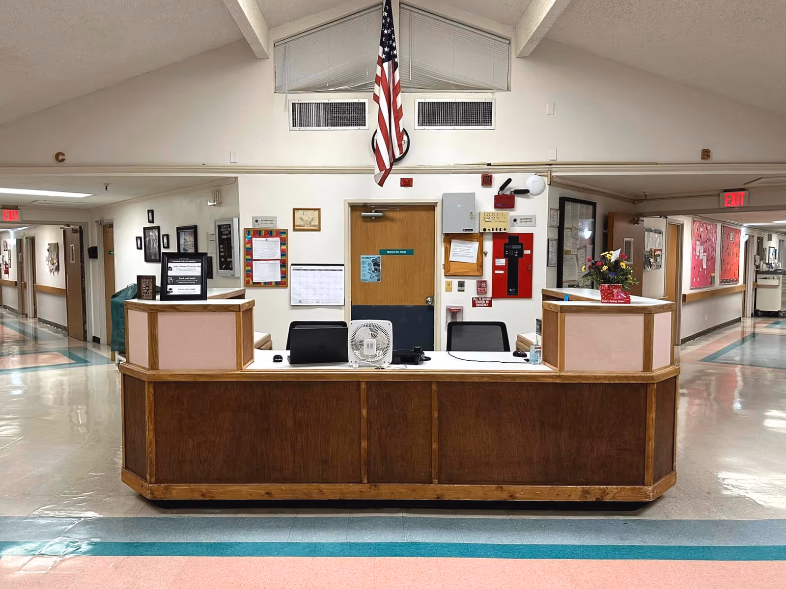 Reception desk area inside a senior living facility with an American flag hanging above a door behind the desk. The desk has a laptop, a small fan, and a flower arrangement. Hallways extend to the left and right with various framed pictures and bulletin boards on the walls.