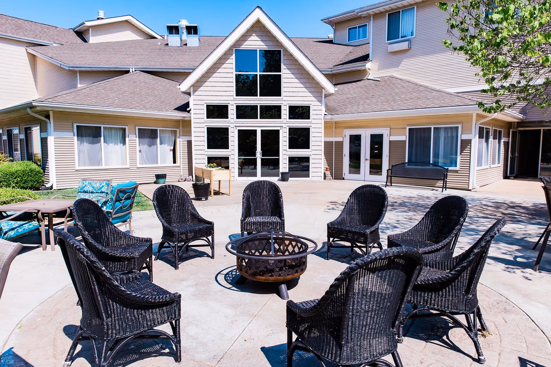Outdoor patio area at College View Manor with a circular arrangement of black wicker chairs around a metal fire pit. The building exterior features beige siding, large windows, and glass doors. There are additional seating areas with blue cushions and a tree providing some shade.