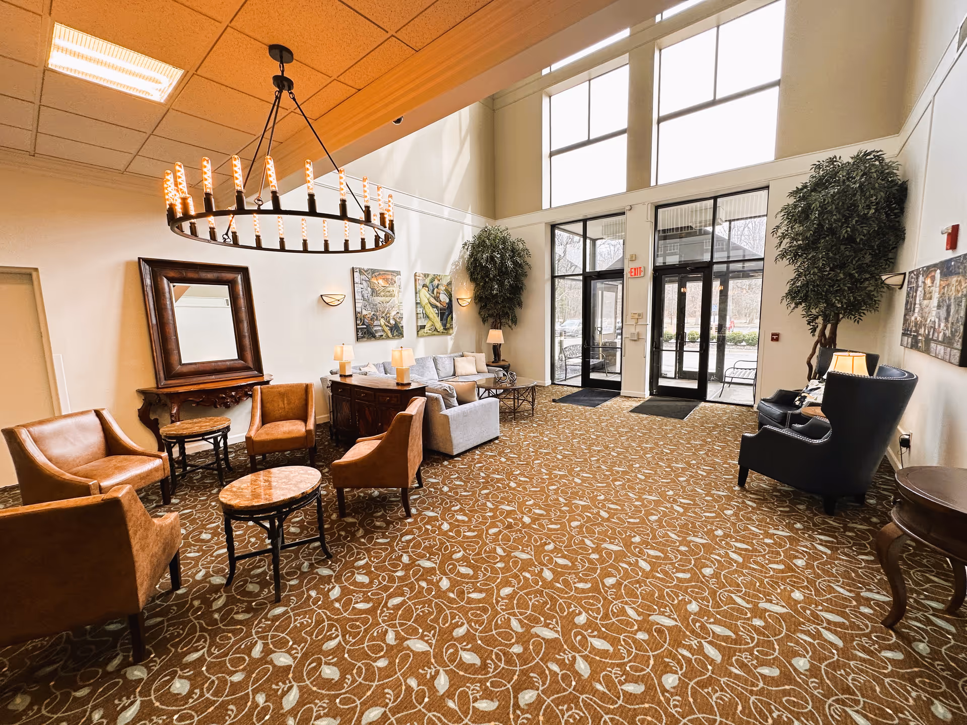A spacious and well-lit lounge area with high ceilings and large windows. The room features a patterned carpet, several brown leather chairs arranged around small tables, a gray sofa with cushions, two large potted plants, wall art, and a large circular chandelier hanging from the ceiling. Glass doors lead outside, allowing natural light to fill the space.