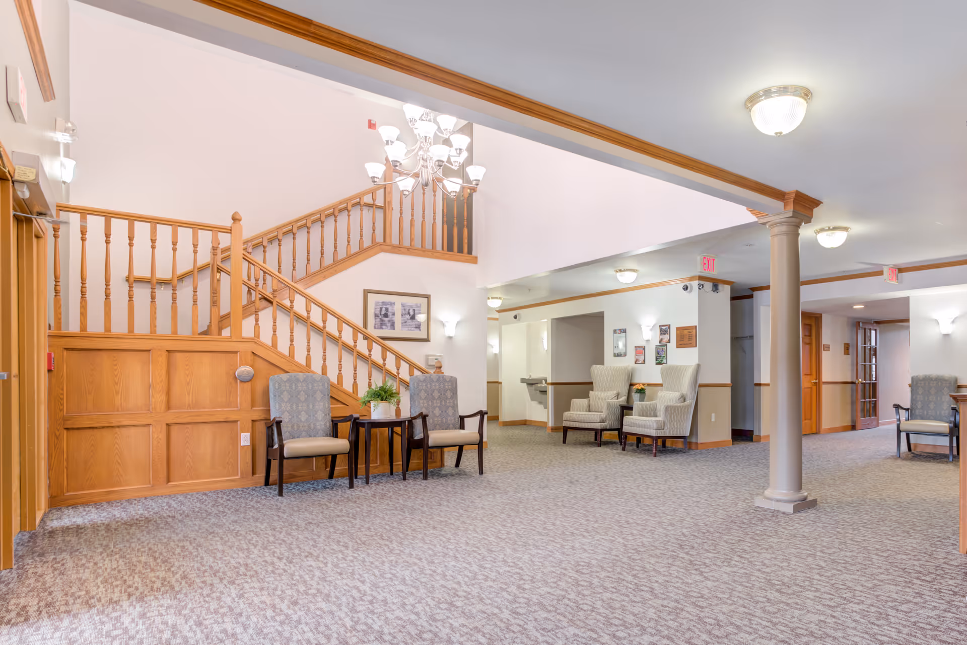 Spacious and well-lit interior common area of a senior living facility featuring a carpeted floor, wooden staircase with railing, multiple upholstered chairs arranged in small seating groups, a chandelier, and ceiling lights. The walls are painted white with wooden trim accents.