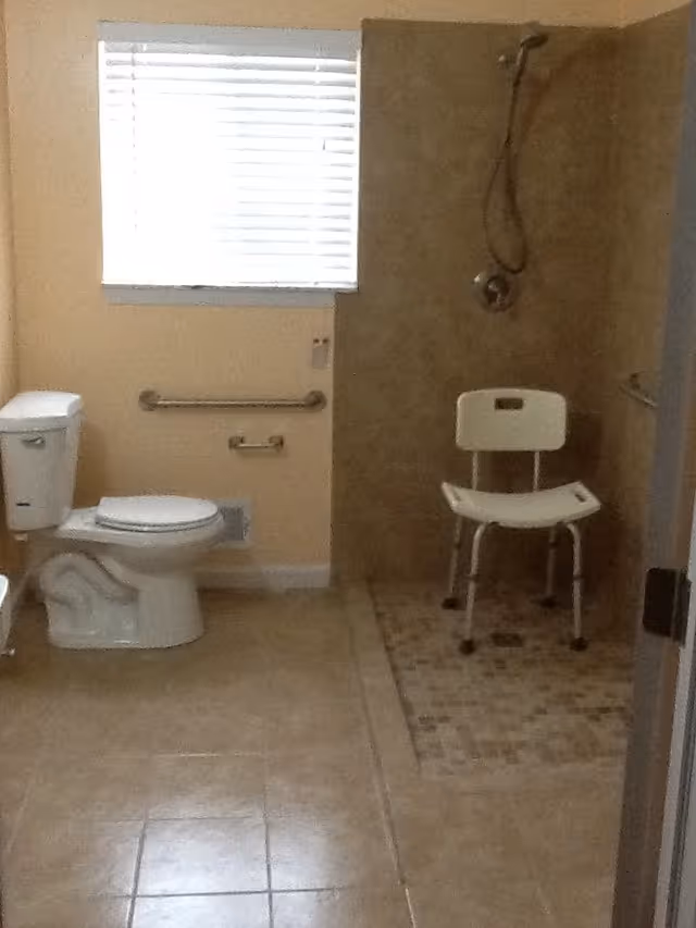 A bathroom with beige tiled floor and walls. On the left side, there is a white toilet with a grab bar mounted on the wall behind it. A window with white blinds is above the toilet. On the right side, there is a walk-in shower area with a handheld showerhead and a white shower chair.