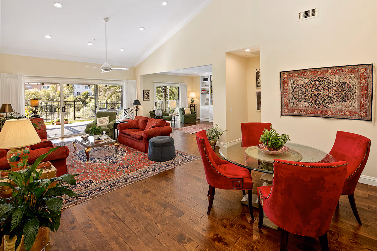 Bright open living room and dining area with red upholstered chairs around a glass table, red sofas, a large patterned rug, and sliding glass doors to a patio.