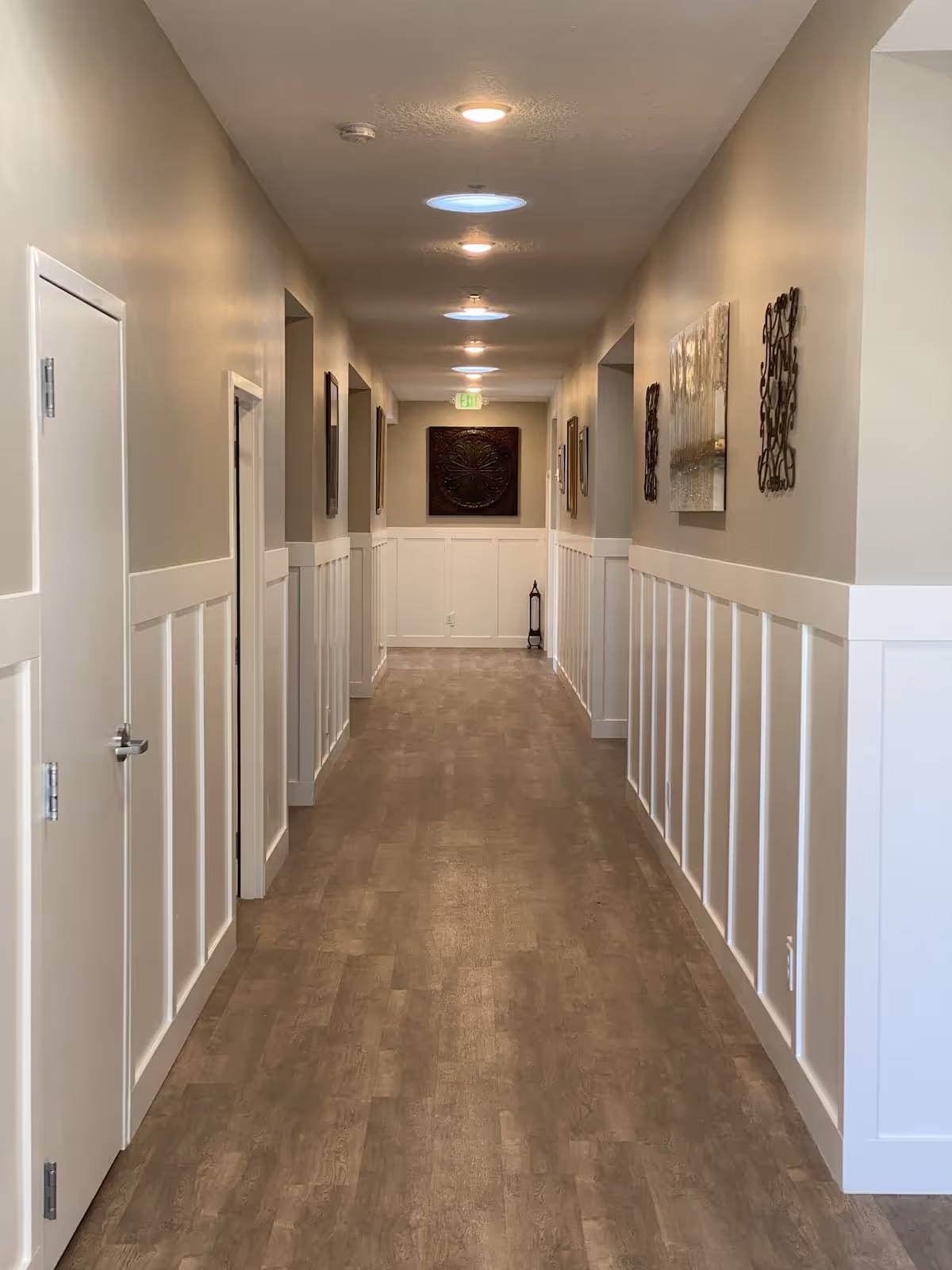 A long, well-lit hallway with wooden flooring and beige walls. The lower half of the walls is white paneling, and there are several framed artworks and decorative pieces hanging on the walls. The ceiling has recessed lights and circular skylights. There are doorways on both sides of the hallway and an exit sign at the far end.