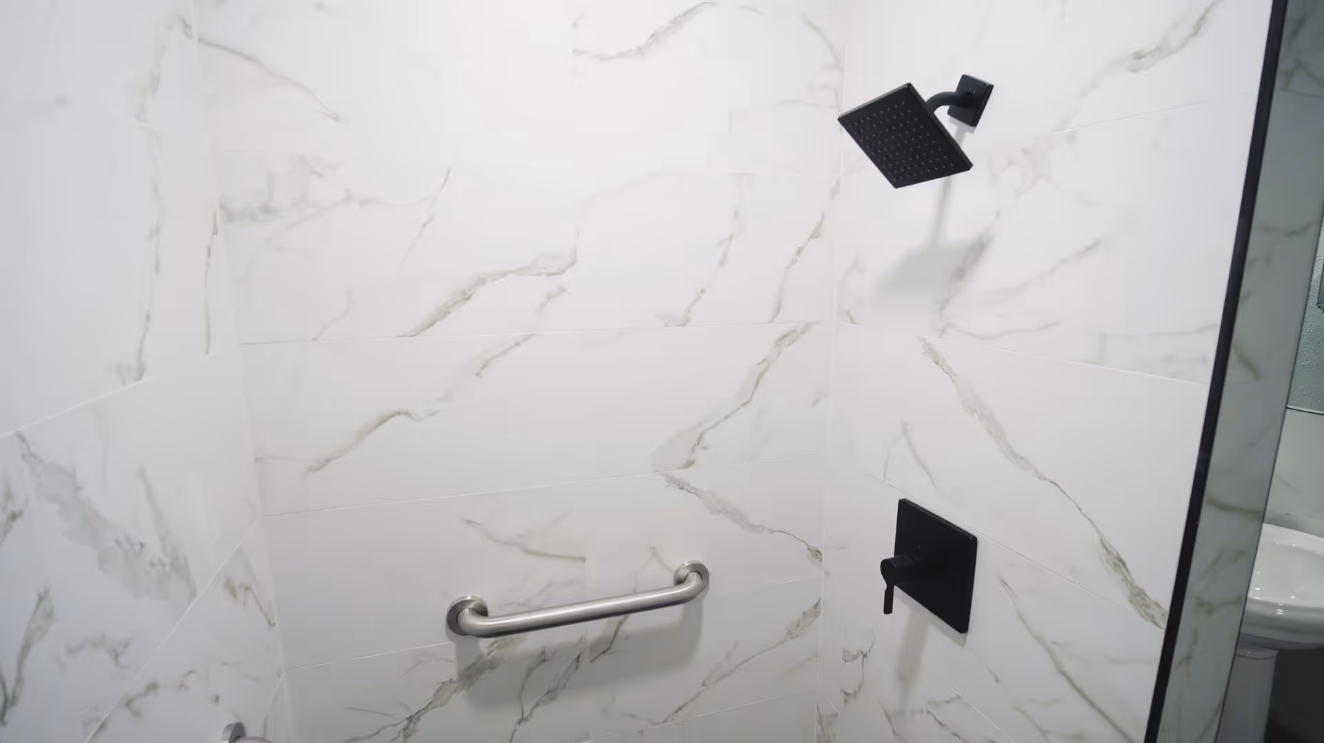 Shower area with white marble-patterned tiles, a black square showerhead, a black control valve, and a horizontal stainless steel grab bar mounted on the wall.