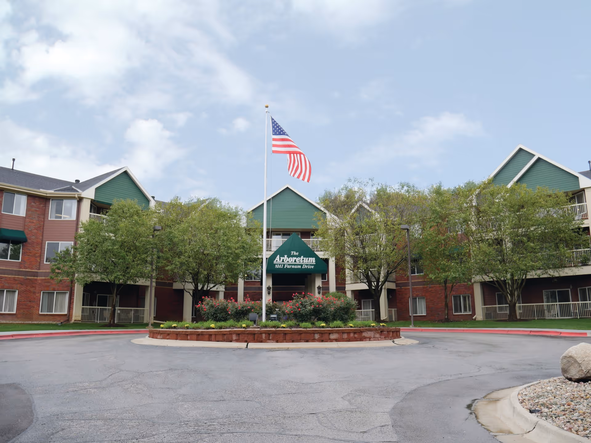 Front exterior view of Immanuel Arboretum Village building with a circular driveway, landscaped garden with flowers and bushes, several trees, and an American flag on a flagpole in the center.