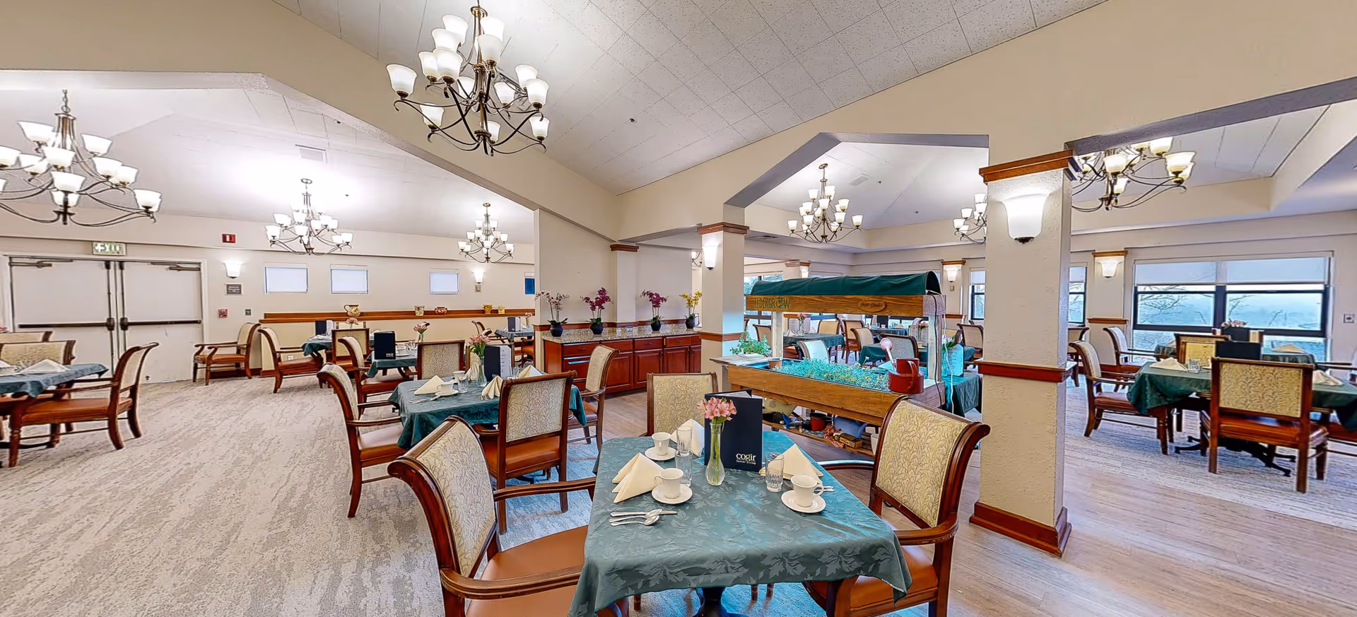 A spacious senior living dining room with multiple tables covered in green tablecloths, each set with cups, glasses, napkins, and silverware. The room features elegant chandeliers hanging from a high ceiling, large windows letting in natural light, and a wooden indoor garden planter in the center. Comfortable upholstered chairs surround the tables, and decorative flowers are placed throughout the room.