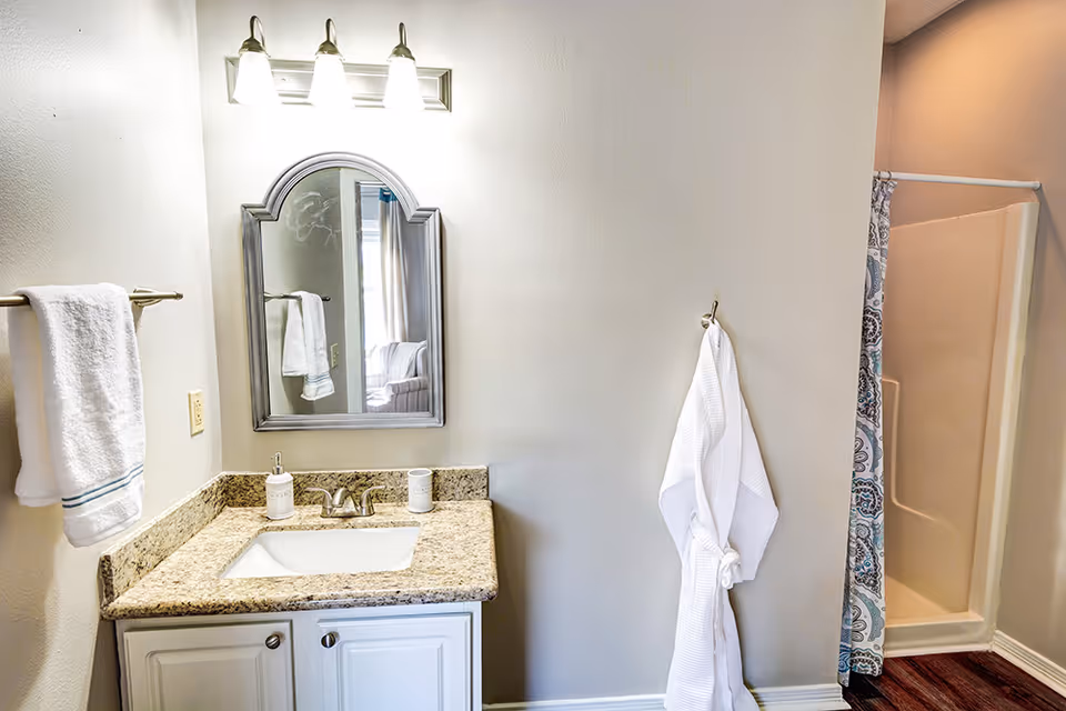 Bathroom with a granite-top vanity, mirror and lights, towels and robe on hooks, and a shower with a patterned curtain.