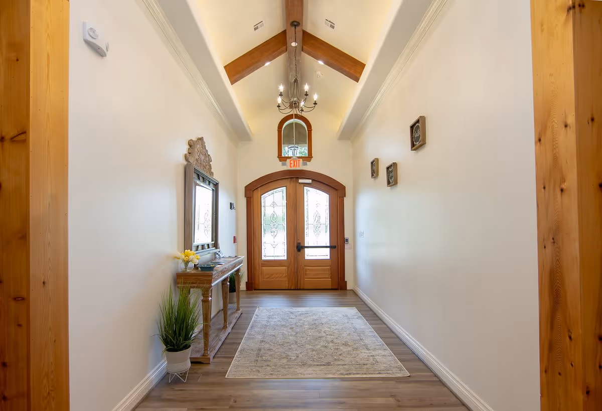 A bright hallway with wooden double doors featuring decorative glass panels at the end. The hallway has light-colored walls, a wooden console table with a mirror above it on the left side, a potted plant on the floor, and three small framed pictures on the right wall. The ceiling is vaulted with exposed wooden beams and a chandelier hanging in the center. A light-colored rug is placed on the wooden floor.