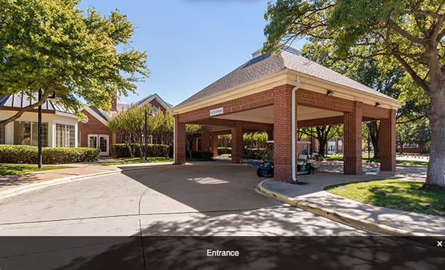 Covered entrance area of a senior living facility named Broadway Cityview with a brick structure supported by columns, surrounded by trees and a driveway.
