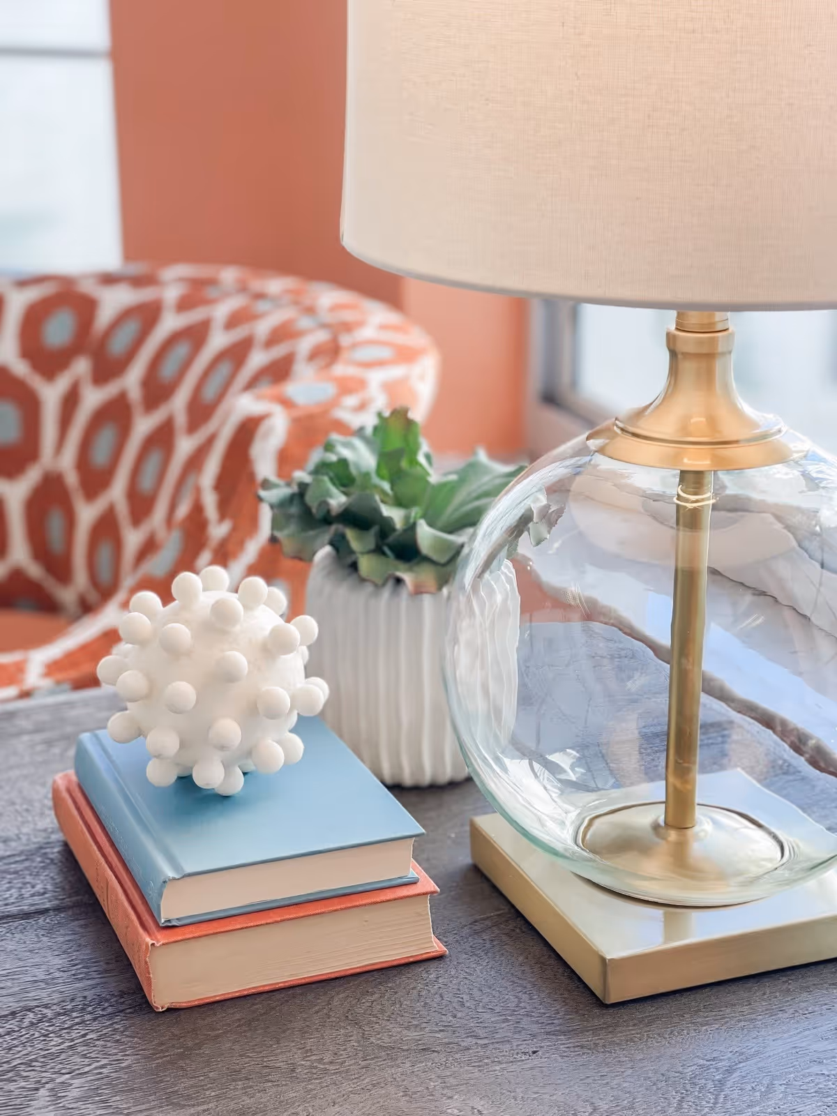 Decorative table vignette with a glass lamp, stacked books, a succulent, and a patterned chair in the background.