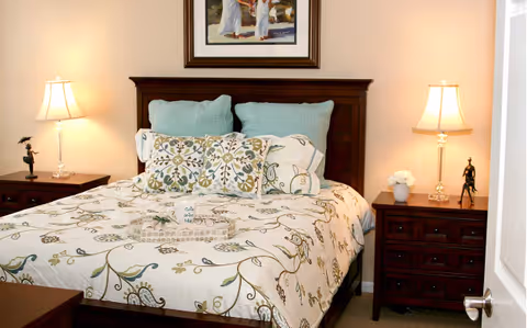 A neatly made bed with a floral patterned comforter and multiple pillows in a bedroom. The bed has a dark wooden headboard. On each side of the bed are matching dark wooden nightstands with table lamps and decorative items. A framed picture hangs on the wall above the bed.