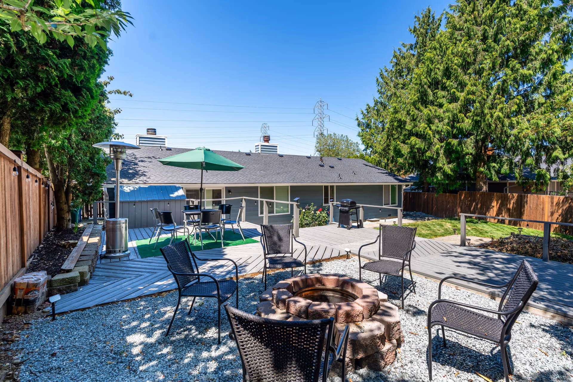 Outdoor patio area with a circular stone fire pit surrounded by six black metal chairs on gravel. Behind the fire pit, there is a wooden deck with a table, four chairs, and a green umbrella. A tall outdoor heater stands near the deck. The area is enclosed by a wooden fence and surrounded by tall green trees under a clear blue sky.