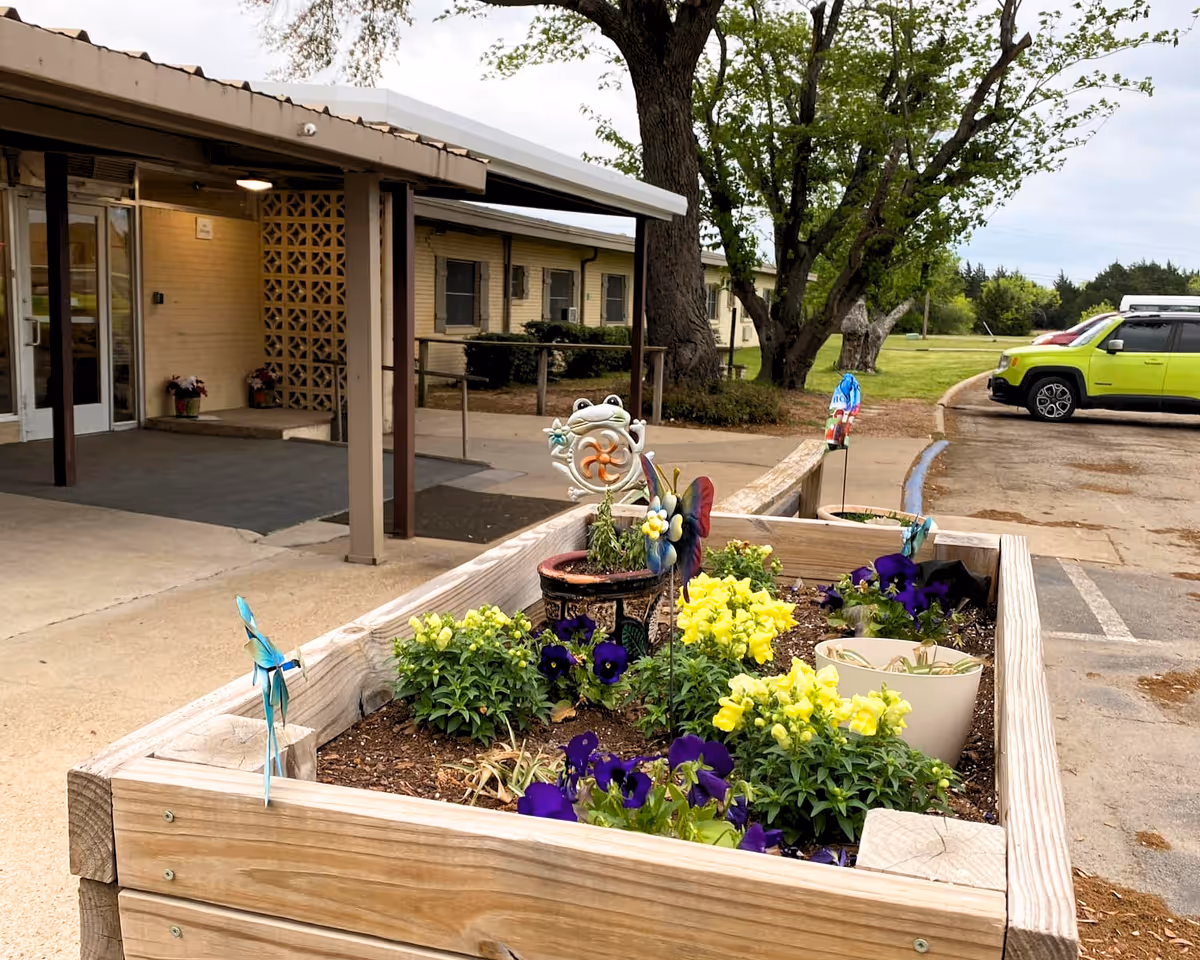 Raised wooden garden bed with yellow and purple flowers and decorative garden stakes in front of the entrance to a single-story beige brick building with a covered walkway. A green car is parked in the background near a grassy area with trees.