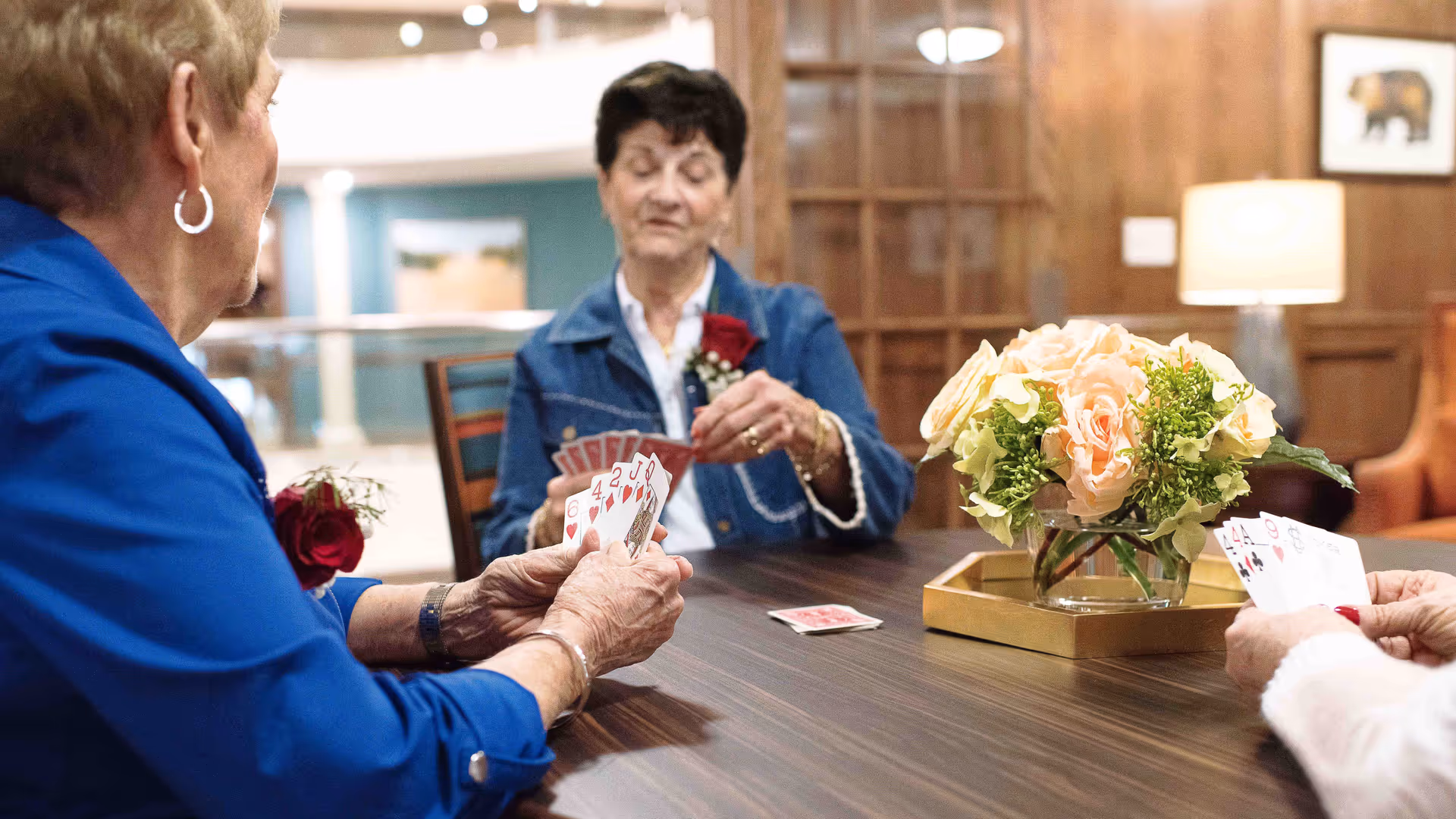 Three elderly women sitting around a wooden table playing cards in a cozy room with wood-paneled walls and a lamp. A vase with a bouquet of light pink and white flowers is placed on the table.