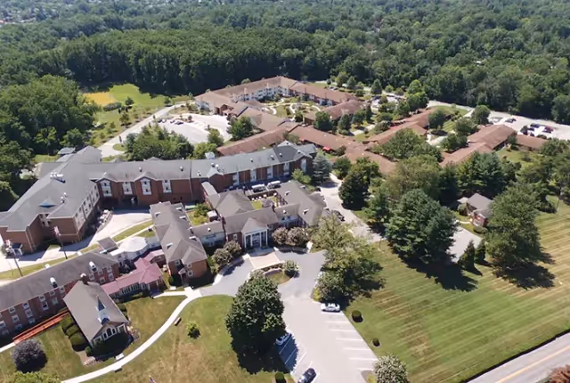 Aerial view of Resorts of Augsburg senior living facility showing multiple connected buildings with brown roofs surrounded by green trees and lawns, parking areas, and a road on the right side.