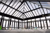 Interior view of a spacious room with a large glass ceiling and walls, allowing natural light to flood the area. The structure features black metal framing and offers a panoramic view of the outside.