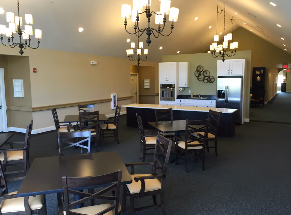 Interior view of a dining area in a senior living facility featuring several dark wooden tables and chairs with light cushions. The room has a high ceiling with multiple chandeliers providing warm lighting. In the background, there is a kitchen area with white cabinets, a stainless steel refrigerator, an oven, and a countertop. The walls are painted beige, and there is a hallway visible to the right.