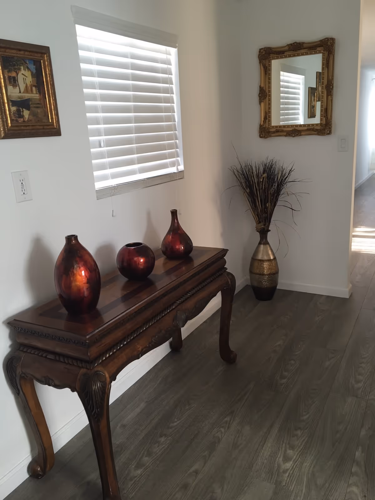 A hallway corner with a wooden console table holding three decorative red vases. Above the table is a window with white blinds and a framed painting on the wall. To the right, a large ornate gold-framed mirror hangs above a tall vase filled with dried decorative grass. The floor is covered with wood-like flooring, and a hallway extends into the background.