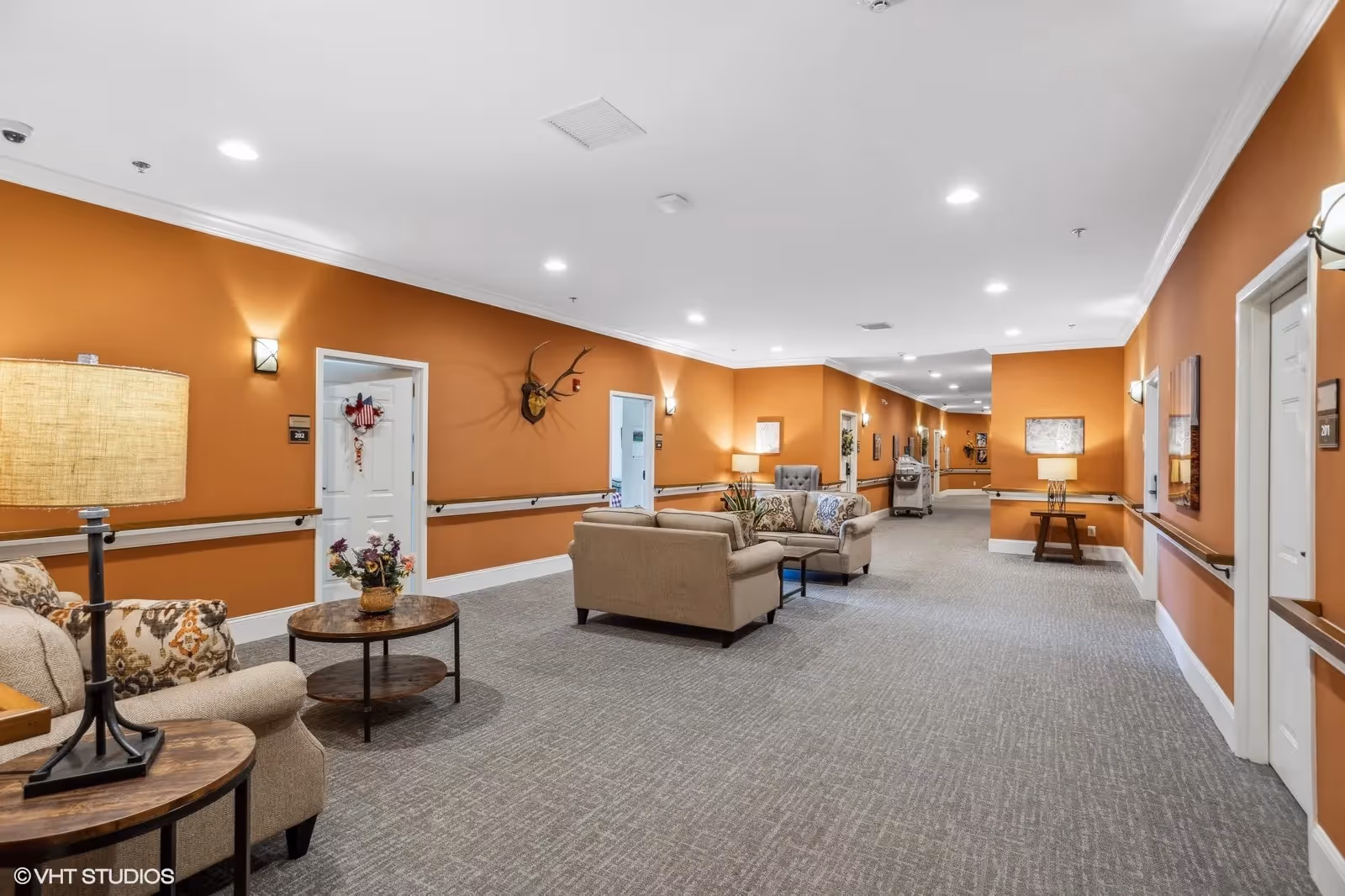 A spacious hallway in a senior living facility with orange walls and gray carpet. The hallway features comfortable seating areas with beige sofas and armchairs, wooden side tables with lamps, and decorative wall art. Handrails run along the walls, and several white doors lead to rooms. The ceiling has recessed lighting.