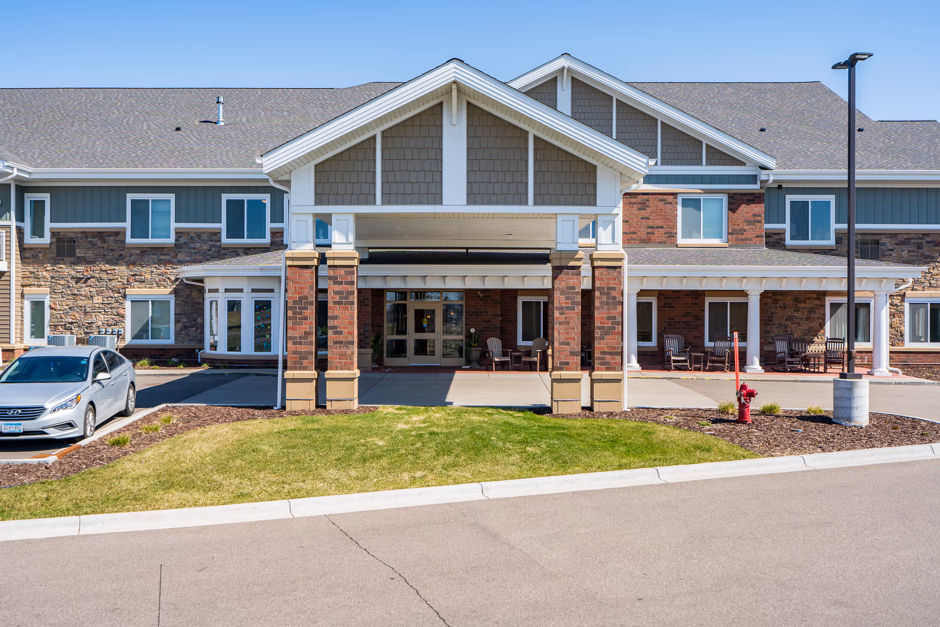 Front exterior view of a senior living facility with a covered entrance supported by brick and white columns. There are several windows, a parked silver car to the left, a fire hydrant on the right, and a well-maintained lawn and driveway in front.