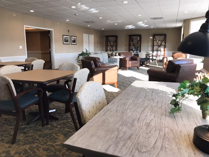 A well-lit common area with several tables and chairs in the foreground and multiple armchairs and sofas arranged in seating groups in the background. The room has beige walls, carpeted floors, and large windows letting in natural light. There are decorative plants and framed pictures on the walls.