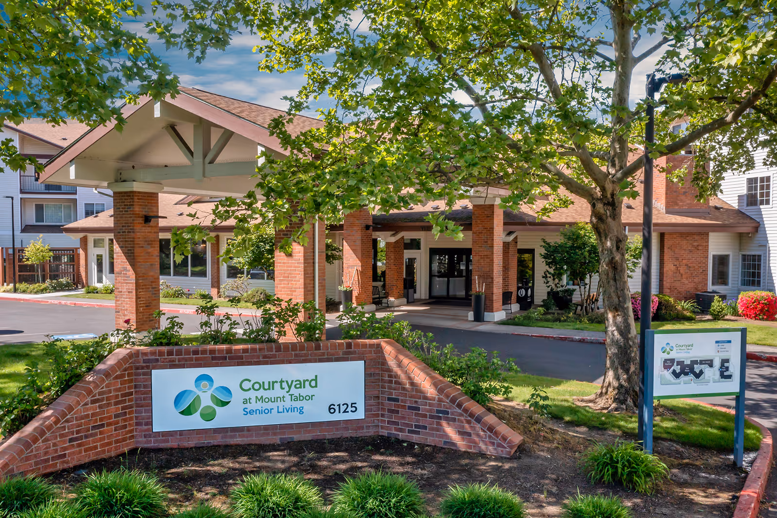 Exterior view of Courtyard at Mount Tabor Senior Living facility showing the entrance with a covered drop-off area supported by brick columns, surrounded by trees and landscaping. A brick sign with the facility name and address is visible in the foreground.