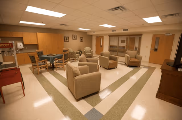 A spacious common area with beige armchairs arranged in a circle, a table with four chairs, wooden cabinets along the wall, a popcorn machine, and a television on a wooden stand. The room has a tiled floor with a geometric pattern and fluorescent ceiling lights.