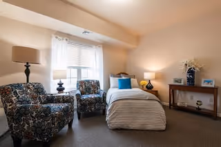 A cozy bedroom with a single bed covered in a beige quilt and a blue pillow. Two patterned armchairs are positioned near a window with white curtains. The room also features a wooden console table with decorative items and two table lamps providing warm lighting.