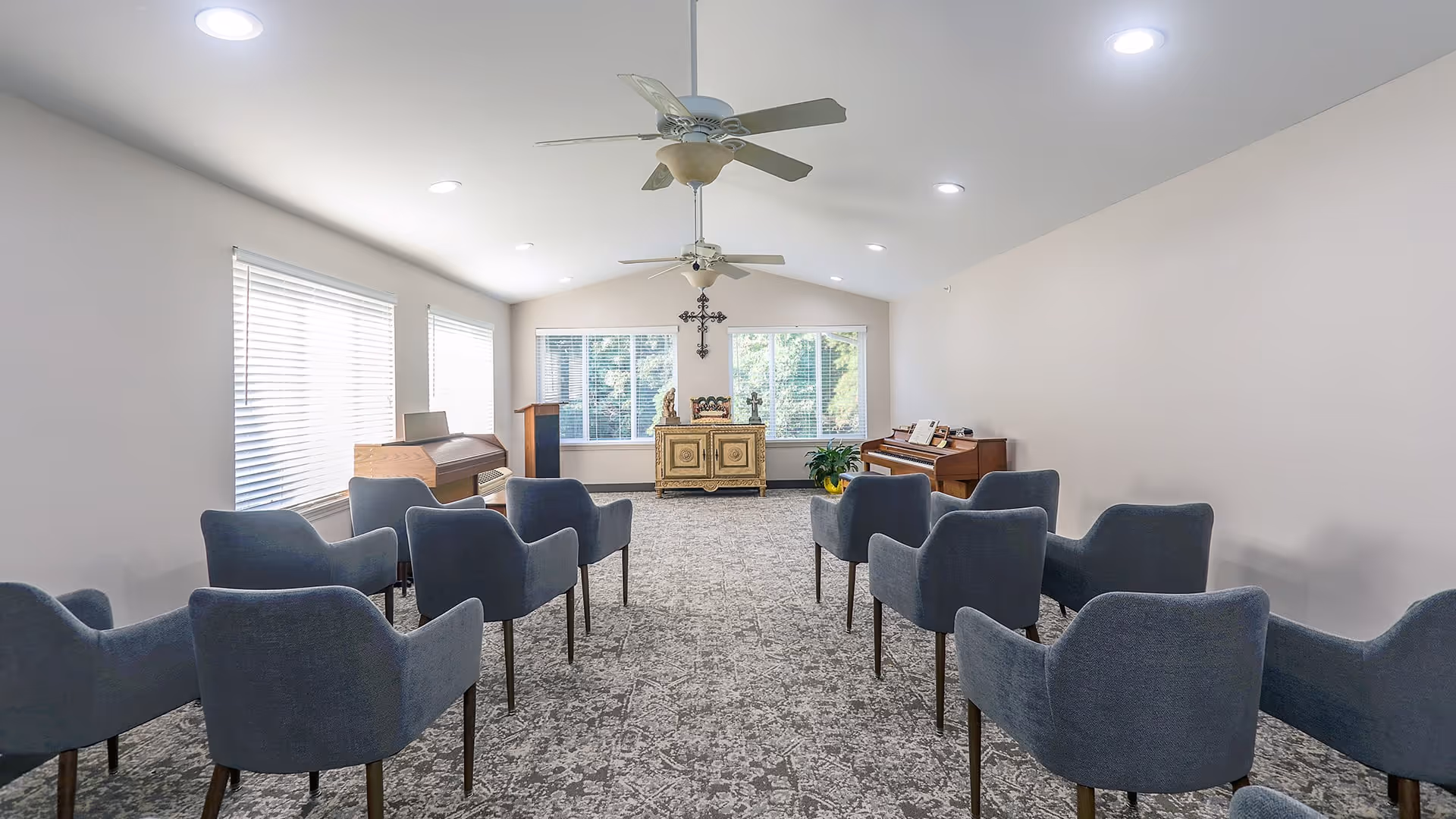 A bright, carpeted room with multiple blue upholstered chairs arranged in rows facing a decorative cabinet and two pianos near large windows. The room has white walls, ceiling fans, and recessed lighting.