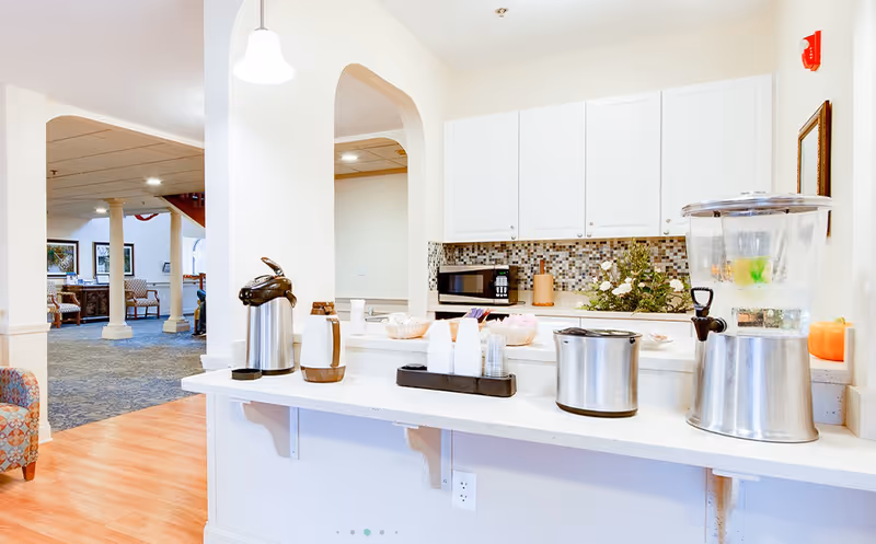 A bright and clean kitchenette area with white cabinets, a mosaic tile backsplash, and a countertop holding a water dispenser, coffee thermos, cups, and other beverage supplies. In the background, there is a seating area with chairs and columns, and a carpeted floor.