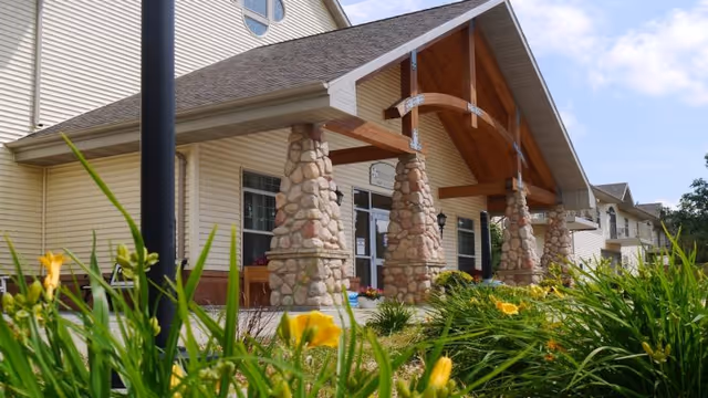 Entrance of a senior living facility with a covered porch supported by stone pillars, surrounded by green plants and yellow flowers under a clear blue sky.