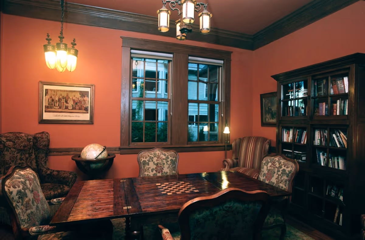 A cozy room with warm orange walls featuring a wooden table with a built-in chessboard, surrounded by upholstered chairs with floral patterns. There is a globe on a stand near the window, a wooden bookshelf filled with books, and two armchairs with different upholstery patterns. The room has wooden window frames and two hanging light fixtures.