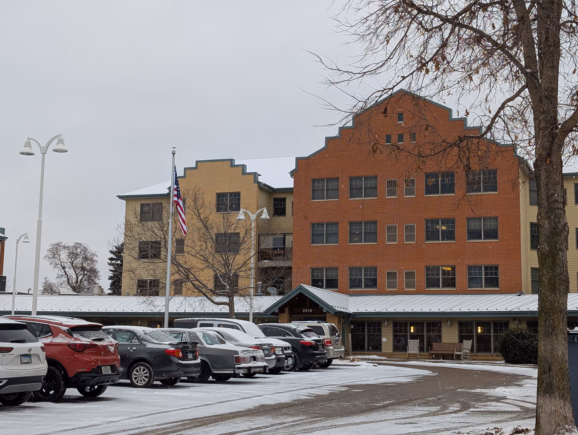 Exterior view of a multi-story senior living facility building with a mix of red brick and beige siding. There is a parking lot in front with several cars covered lightly with snow. An American flag is flying on a flagpole near the entrance. Leafless trees and snow-covered ground indicate a winter setting.