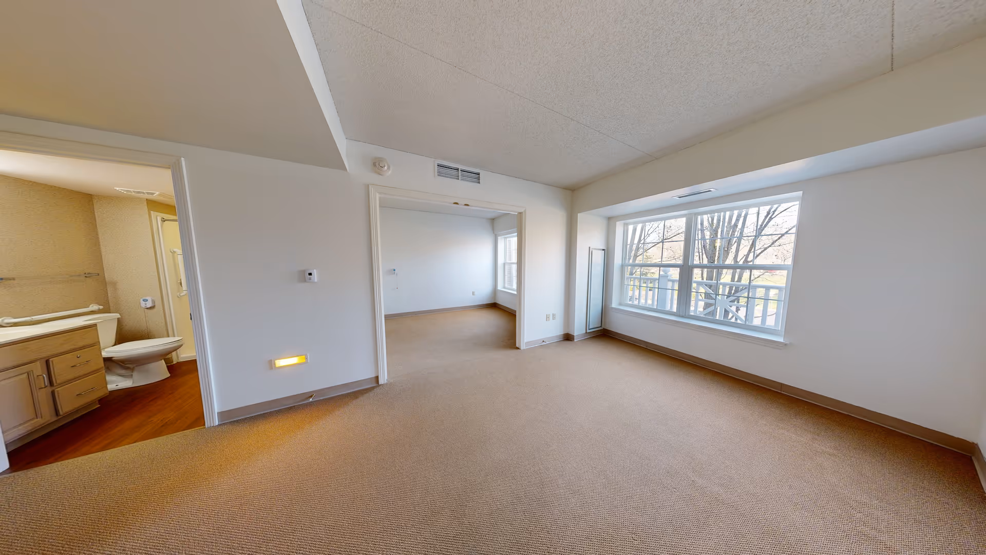 Empty senior living apartment with beige carpet, white walls, a large window with a view of trees, an open doorway leading to another room, and a bathroom visible on the left side with a toilet and vanity.