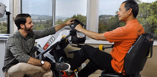 A man in an orange shirt is using a SciFit exercise machine while another man, wearing a plaid shirt and beige pants, kneels beside him, assisting or encouraging him. They are in a room with large windows showing a scenic outdoor view with trees and hills.