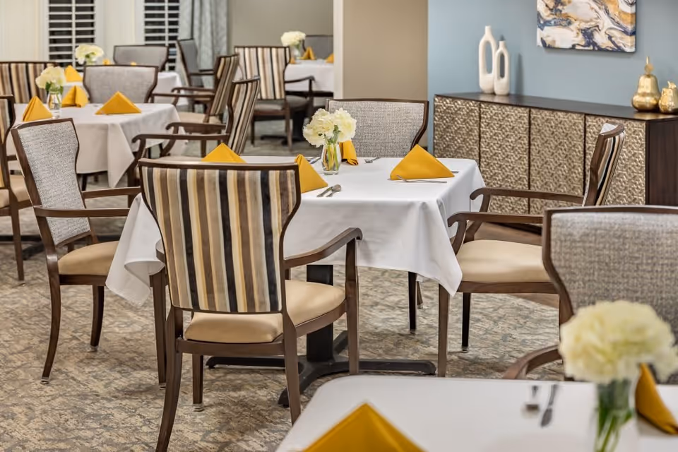A dining room with tables covered in white tablecloths, each set with yellow folded napkins and a small vase of white flowers. The room has cushioned chairs with striped and solid upholstery, a patterned carpet, and a sideboard with decorative vases and artwork on the wall.