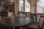 Interior view of a common area in a senior living facility with a round dark wooden table surrounded by four upholstered chairs. In the background, there are large windows with beige curtains, a patterned armchair, and a side table with a decorative lantern. The room has neutral-colored walls and carpeted flooring.