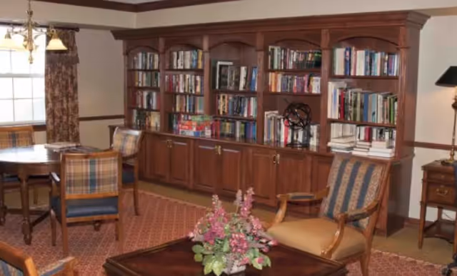 A cozy living room area with a large wooden bookshelf filled with books, a round wooden table with four plaid upholstered chairs, a wooden armchair with striped upholstery, a wooden coffee table with a floral arrangement, and a window with patterned curtains.