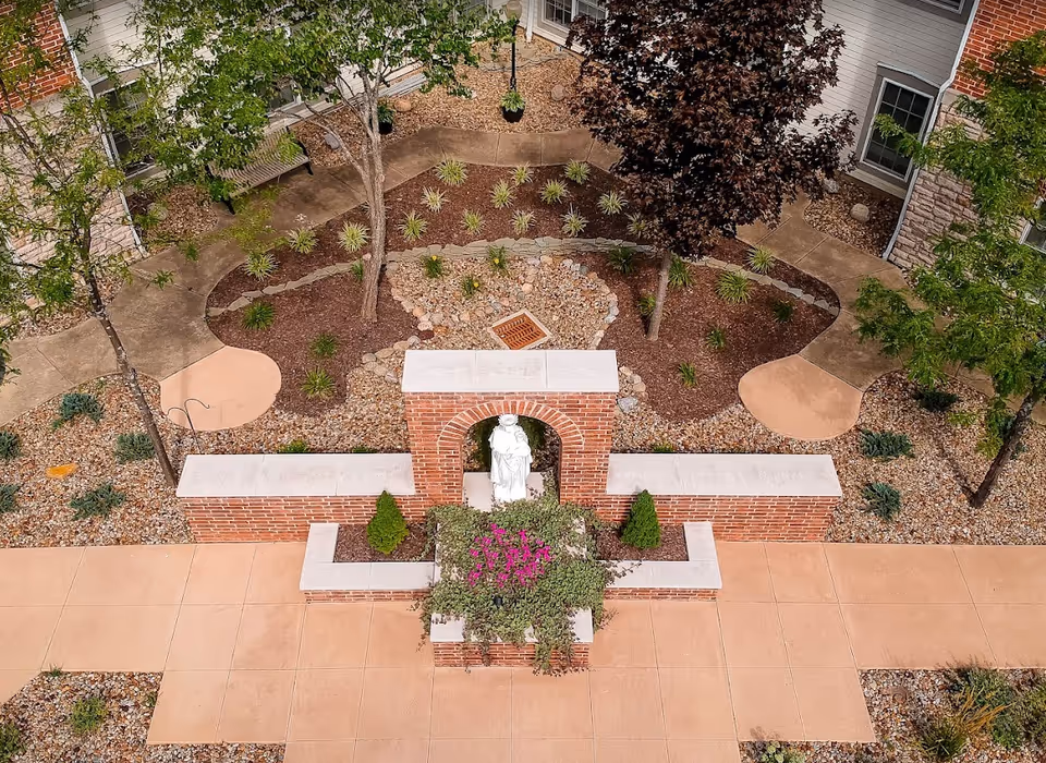 A landscaped outdoor courtyard area with a brick archway featuring a white statue underneath. The courtyard has paved walkways, small trees, shrubs, and decorative rocks. The surrounding building walls have windows and a mix of brick and siding.