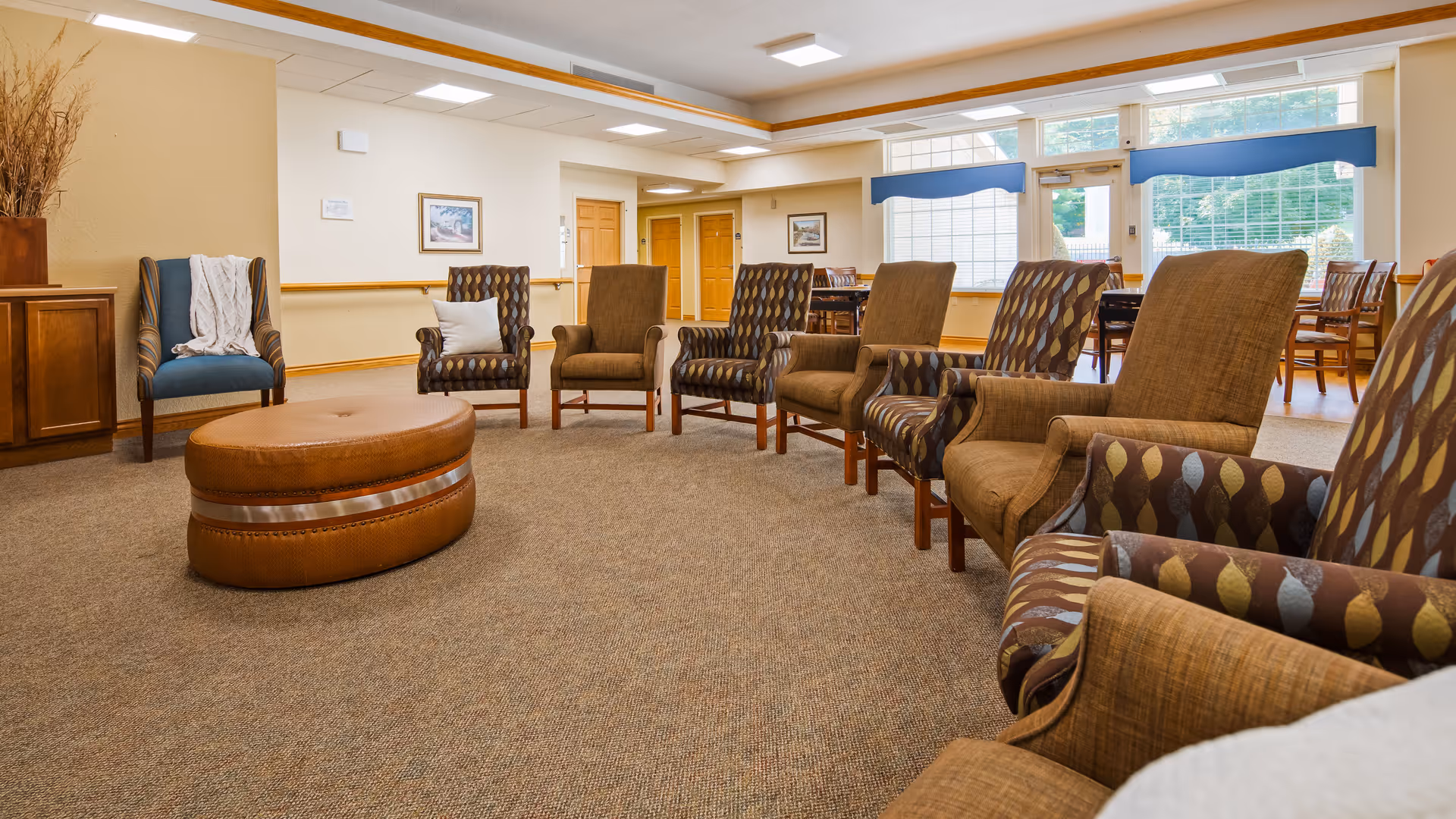 A spacious senior living facility common area with a circle of upholstered armchairs surrounding a large round leather ottoman. The room has beige walls, carpeted floor, large windows with blue valances, and wooden furniture including a cabinet and dining tables with chairs in the background.